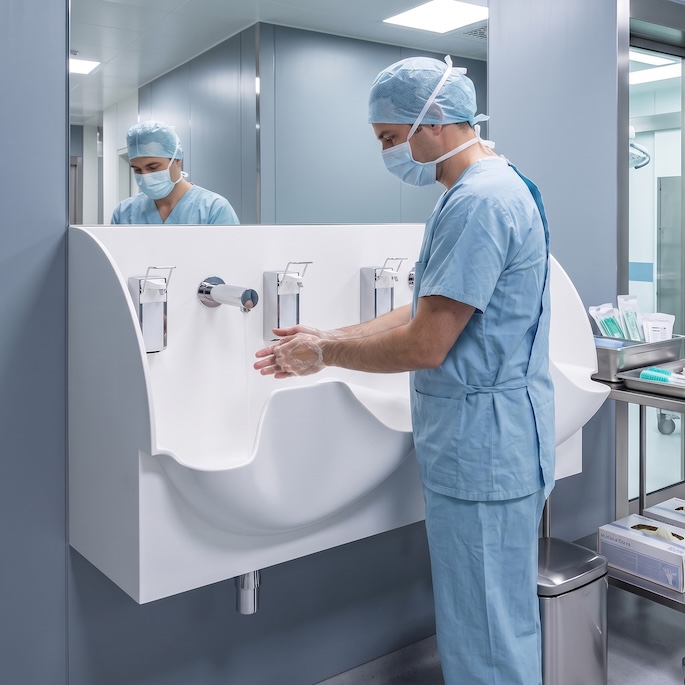A male surgeon in blue scrubs, a surgical mask, and a cap washes his bare hands at a modern, seamless white solid-surface double surgical sink. The room has light grey-blue walls, and a large mirror above the sink reflects the surgeon.