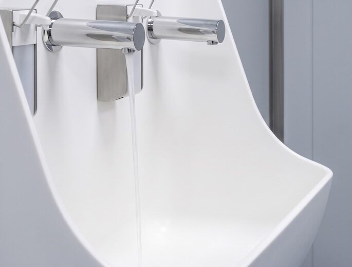 A detailed close-up shot of a seamlessly integrated white Corian Abra solid surface sink unit. Water flows continuously from the left of two wall-mounted polished chrome faucets into the basin. Integrated soap dispensers are visible above the faucets, set against grey wall panels in a sterile clinical environment