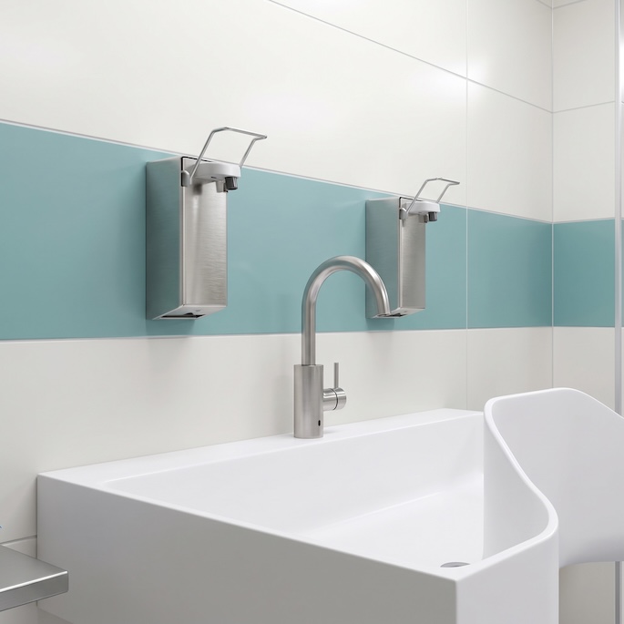A sterile, modern medical hand washing station featuring a large, seamless, white Corian solid surface sink and a single curved stainless steel gooseneck faucet. The background wall is tiled with large white tiles and a central horizontal band of turquoise tiles. Above the sink are two wall-mounted stainless steel soap or sanitizer dispensers.